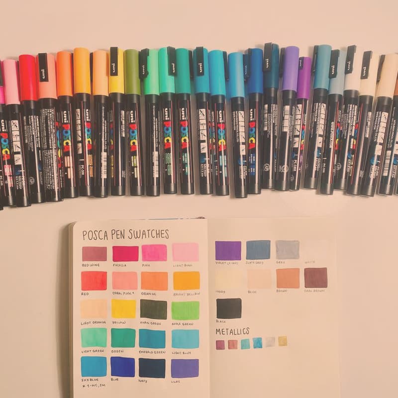 An array of Posca pens above an open sketchbook where I've swatched them out