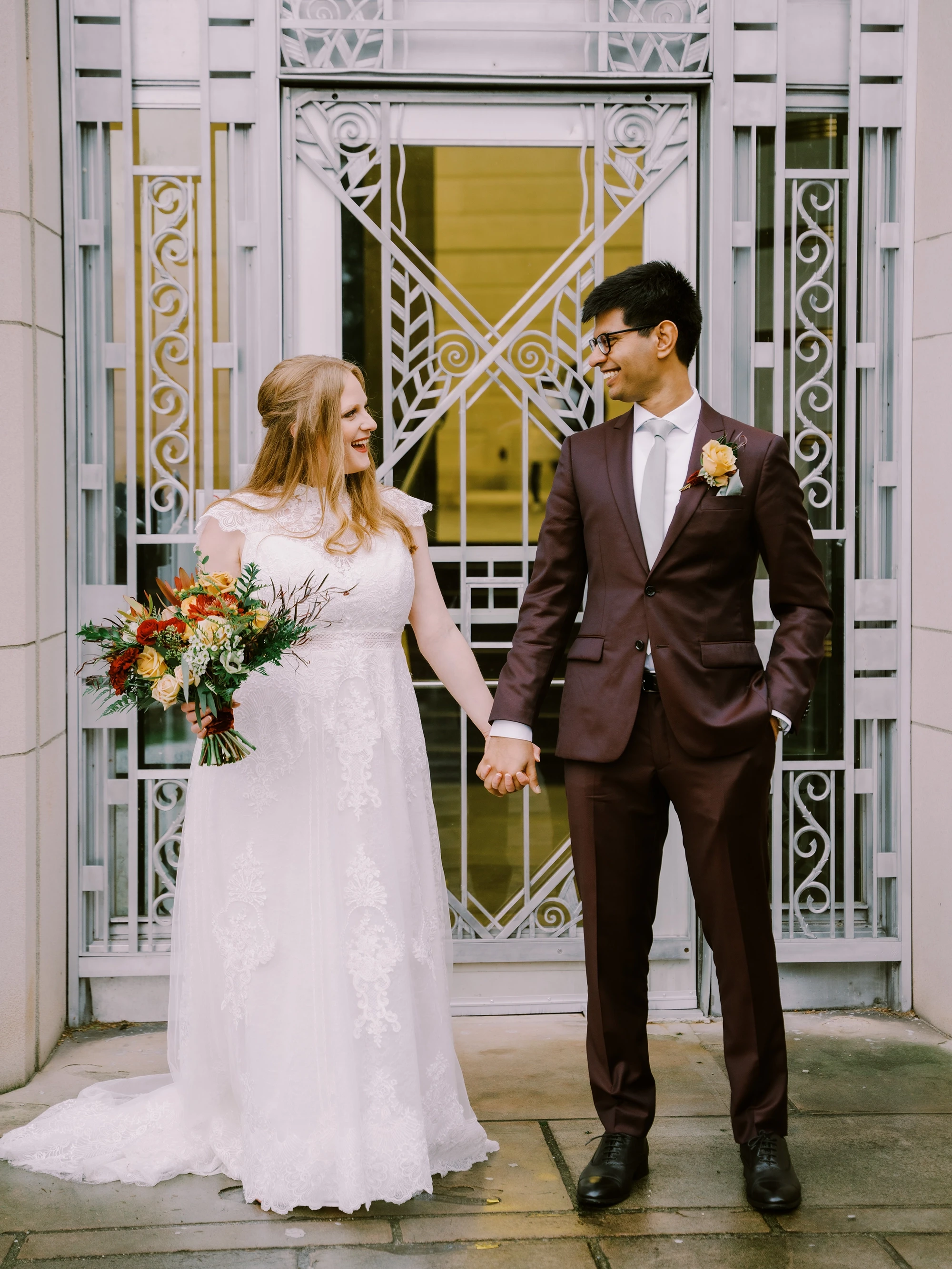 My husband and I grinning at each other in front of art deco metalwork. I’m wearing a classic western wedding dress in white, and he has on a burgundy suit.