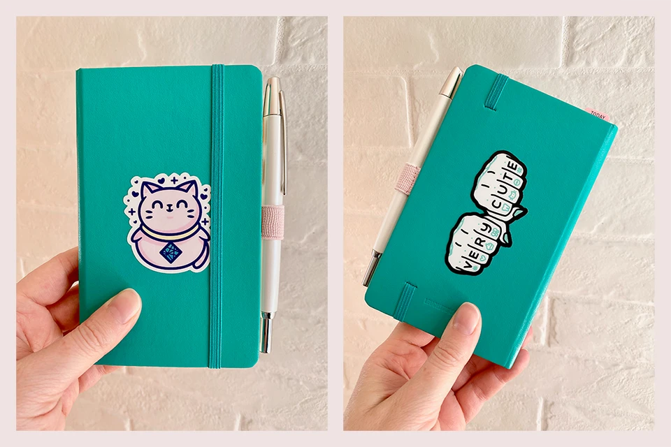 A small teal notebook with a shiny white pen in a light pink pen loop. On the front is a sticker of a chubby pink cat with the old Netlify logo on its collar. The sticker on the back is a drawing of knuckles with very cute tattooed onto them, along with cutesy symbols like a heart, tooth, and cat.
