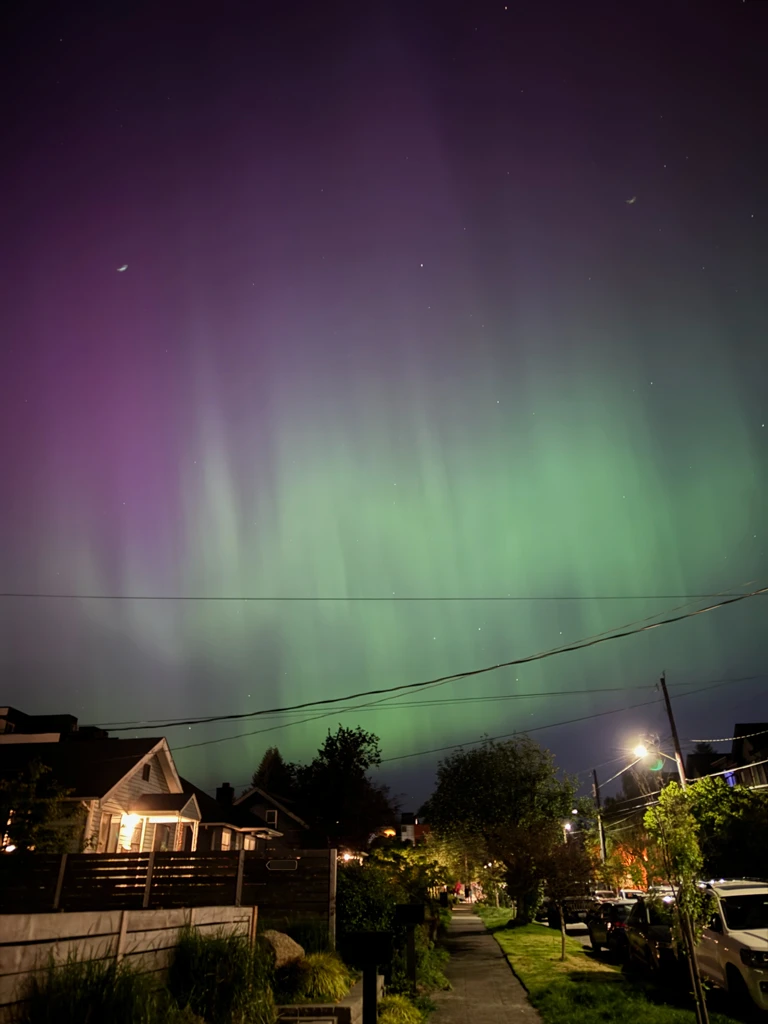 A very strong aurora over a fairly brightly lit residential street. There is a curtain of bright green waves of light below a backdrop of warm violet tones.