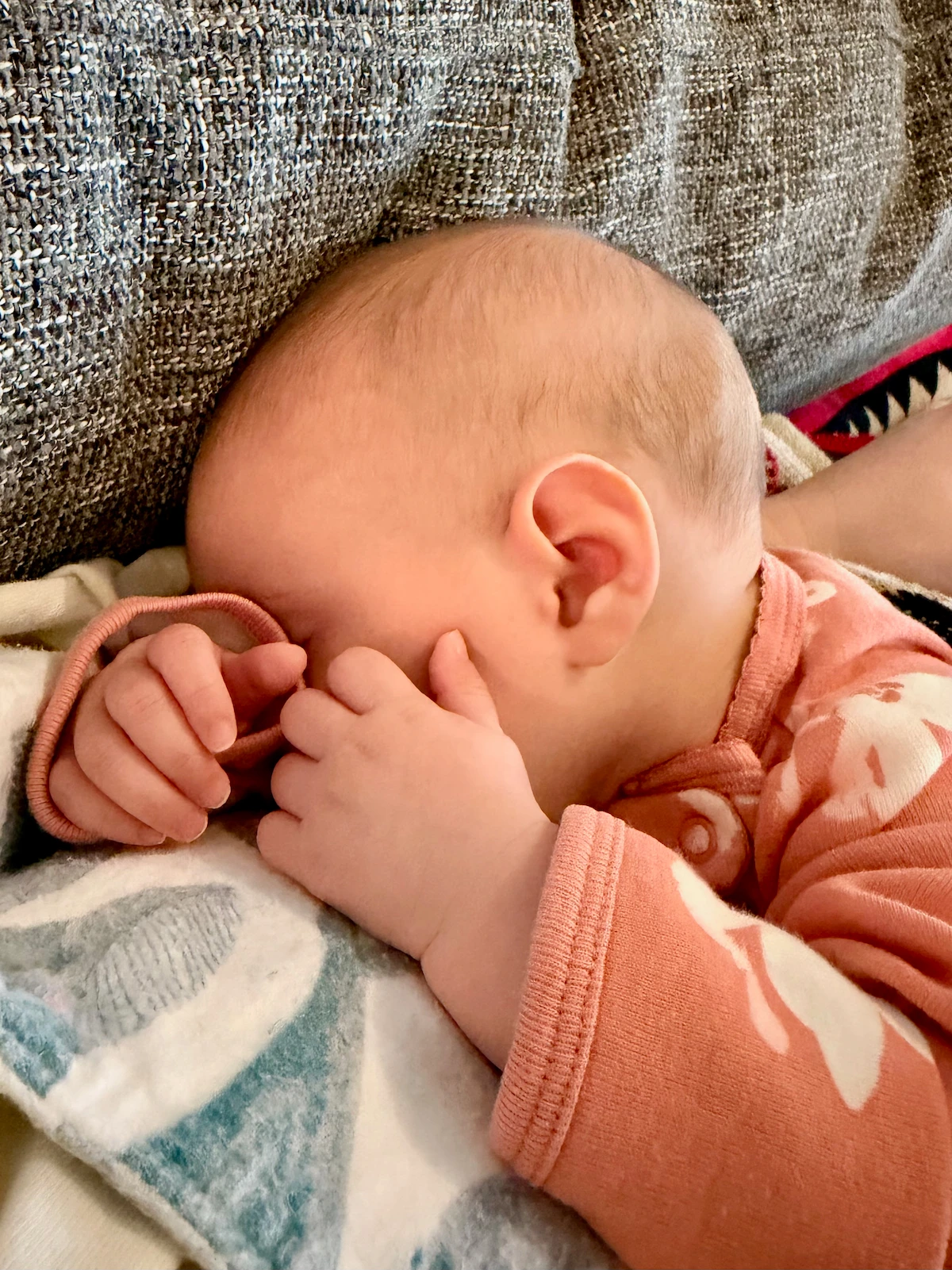 A 3-month baby cozied up on me on a grey couch. She is wearing a pink floral onesie and her chubby little hands cover her face