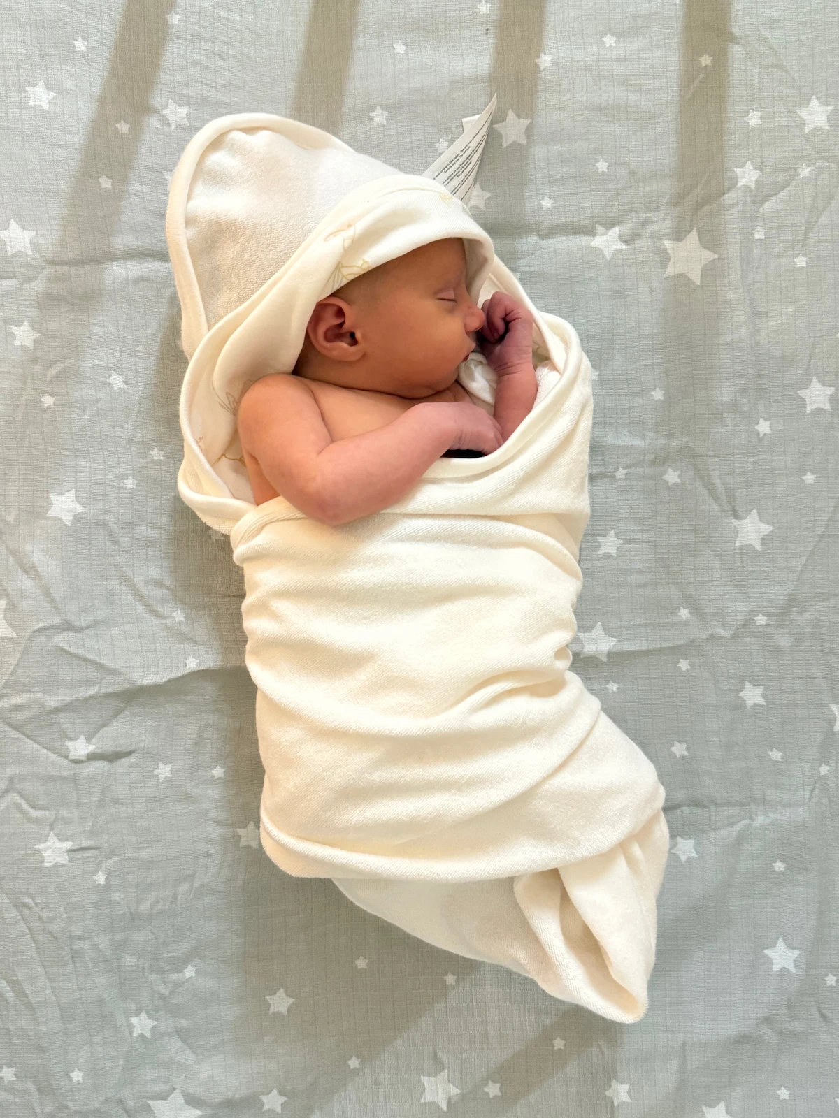 A little baby snuggled up in a hooded towel with her hands curled up next to her face. Light streams into view between the bars of the crib.