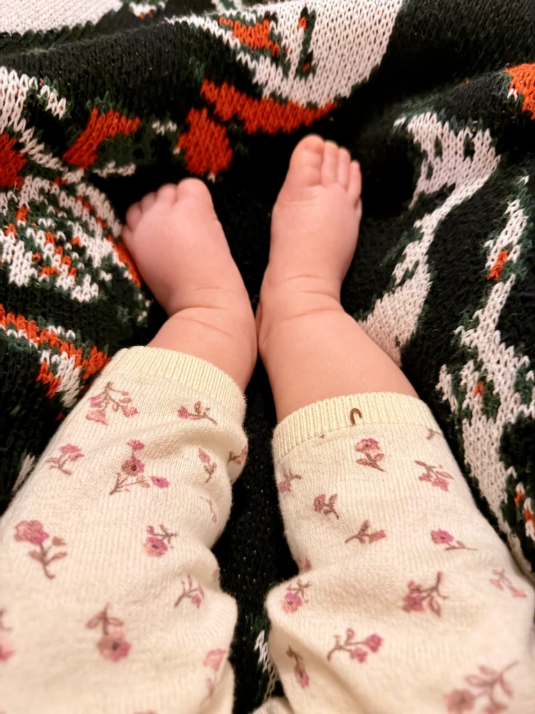 A white-presenting baby's bare little feet cozied up in a knitted blanket with a black, red, green, and white design