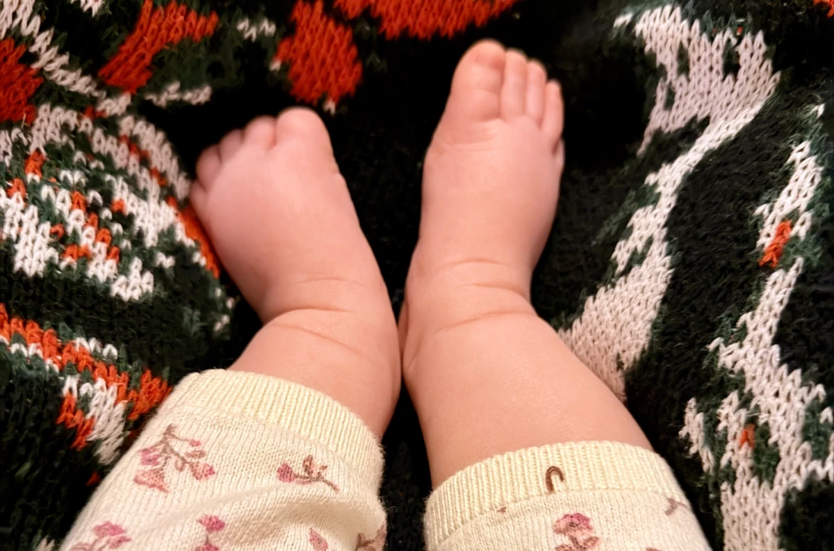 A white-presenting baby's bare little feet cozied up in a knitted blanket with a black, red, green, and white design