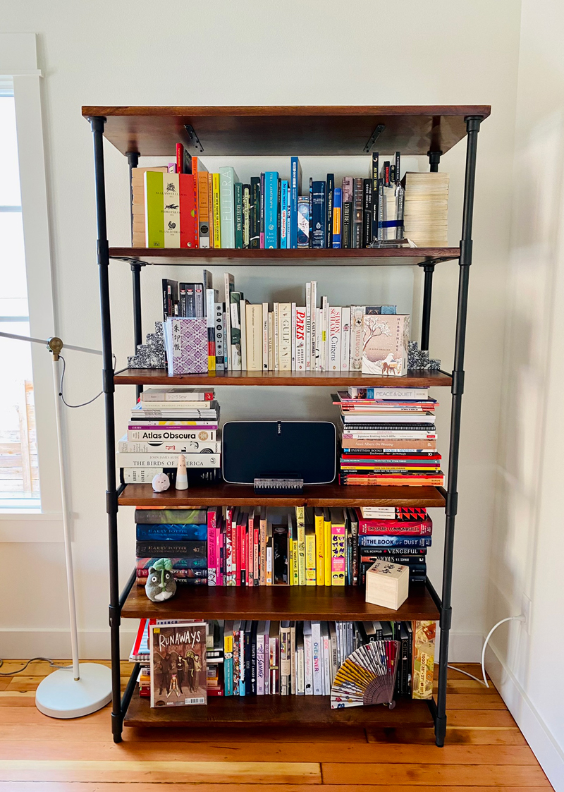 Industrial-style bookcase with five wood shelves and iron piping on the sides. It’s chock full of books of various genres (some sections rainbow ordered), a large Sonos device, and with a couple knickknacks in front of the books.