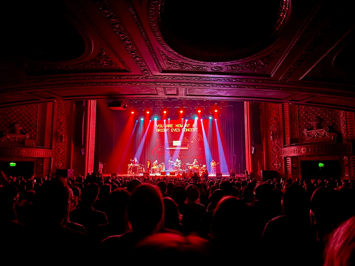 A packed concert hall bathed in red light, with the words 'You are now at a Bright Eyes concert' projected to the back of the stage, while the band plays. The ceiling overhead has a lot ornate, circular crown molding.