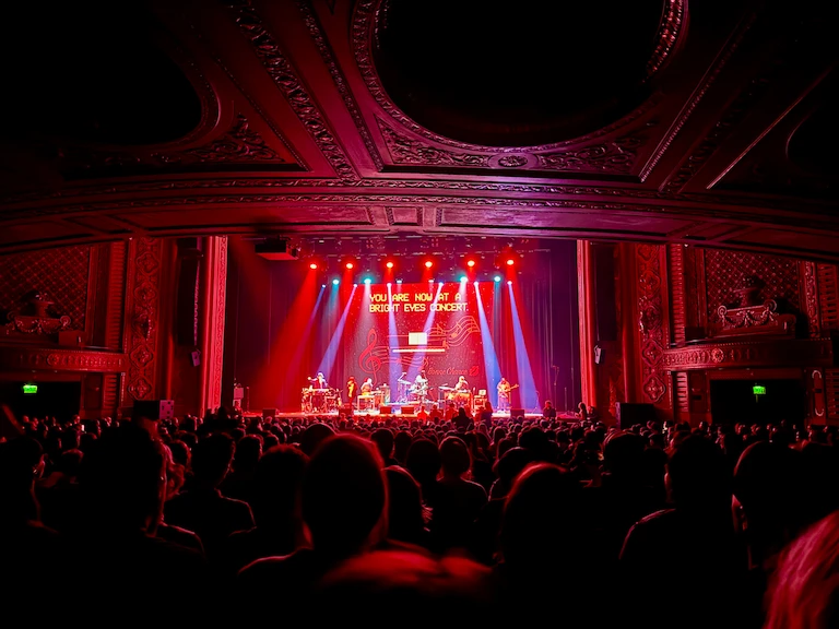 A packed concert hall bathed in red light, with the words 'You are now at a Bright Eyes concert' projected to the back of the stage, while the band plays. The ceiling overhead has a lot ornate, circular crown molding.