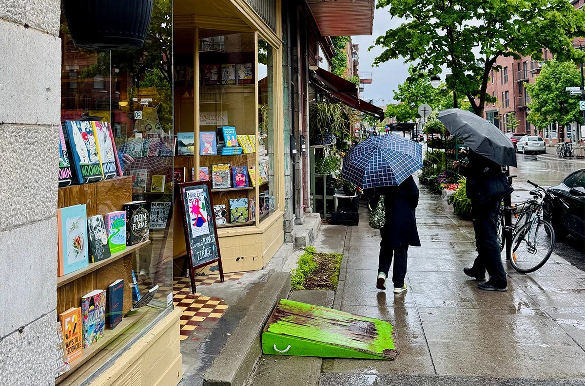 A rainy view of the sidewalk in front of Drawn & Quarterly, a bookstore and publisher specializing in comic books. Two people are walking by with umbrellas.
