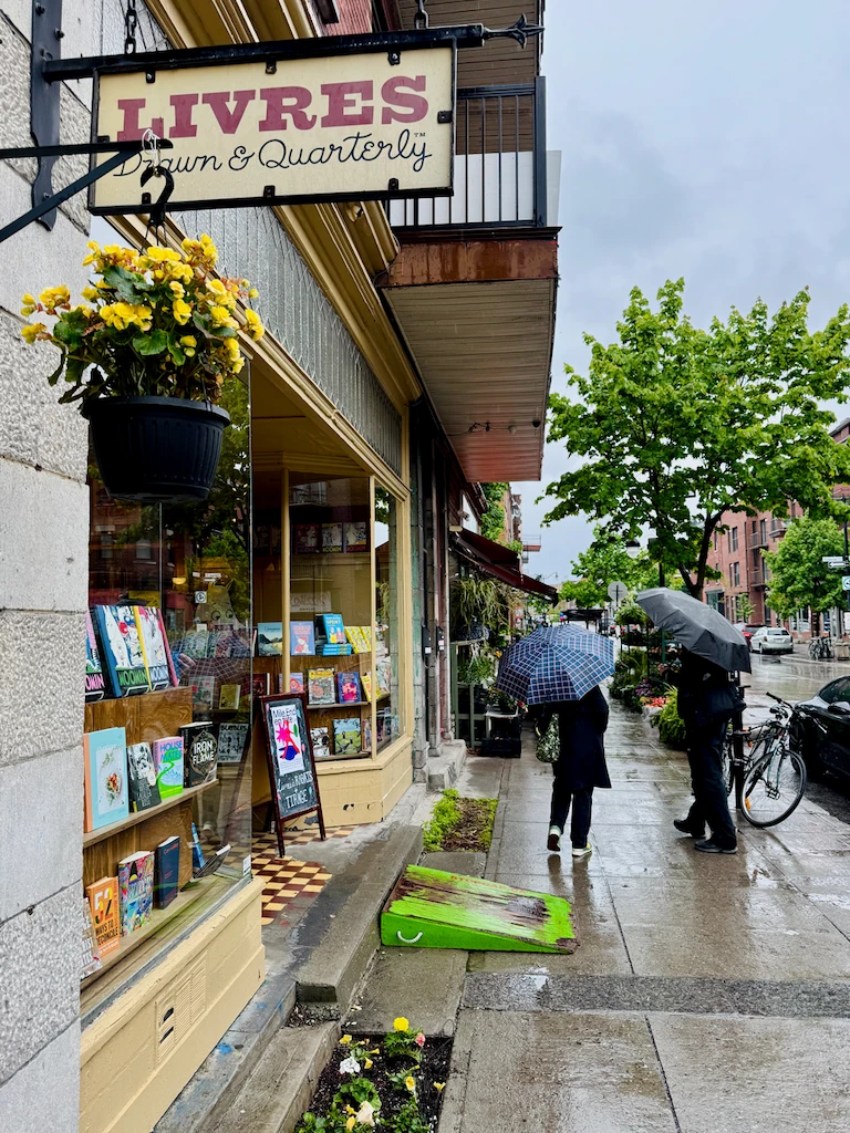 A rainy view of the sidewalk in front of Drawn & Quarterly, a bookstore and publisher specializing in comic books. Two people are walking by with umbrellas, and there's a hanging pot of yellow flowers at the front of the store.