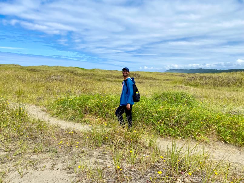 Rahul smiling in a blue hat and jacket on a dirt path in the middle of a bright green field. There are yellow blossoms scattered throughout.