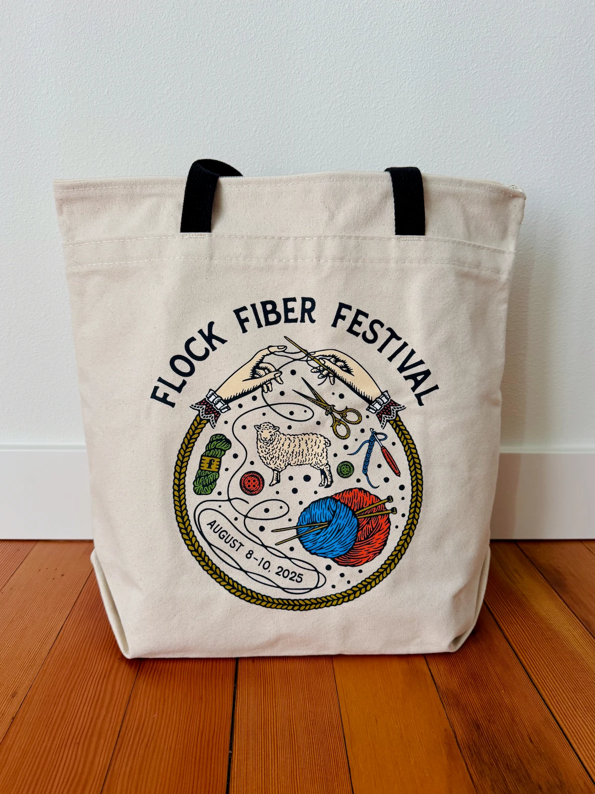 A cream canvas tote bag with a black webbing handle. The Fiber Flock Festival logo is displayed in a large circle on the side.