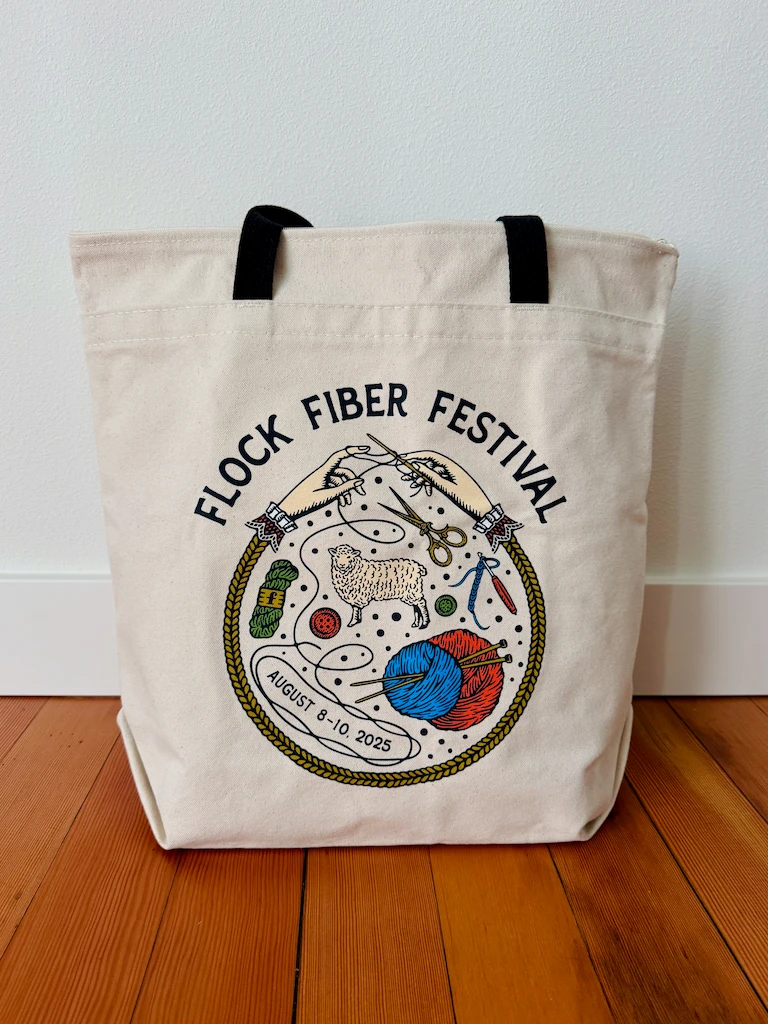 A cream canvas tote bag with a black webbing handle. The Fiber Flock Festival logo is displayed in a large circle on the side.