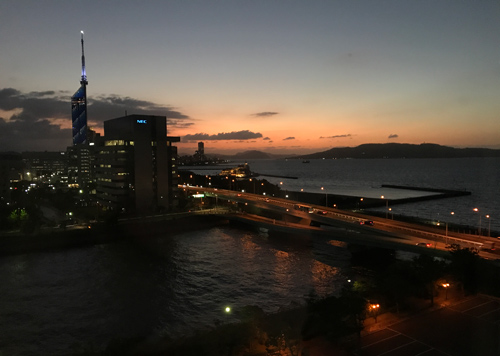 Sunset scene from a high window in Fukuoka, with a couple office buildings, the sea, and distant mountains