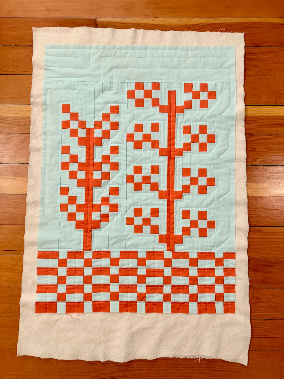 A baby-sized quilt top lying on cotton batting and then a hardwood floor. The foreground includes 2 pixelated plants in a saturated dark orange, and the background is a very light teal blue. The bottom of the quilt shows a checkerboard pattern in the two colors. The quilting traces the outline of the plants, with a gridded quilting pattern for the checkerboard
