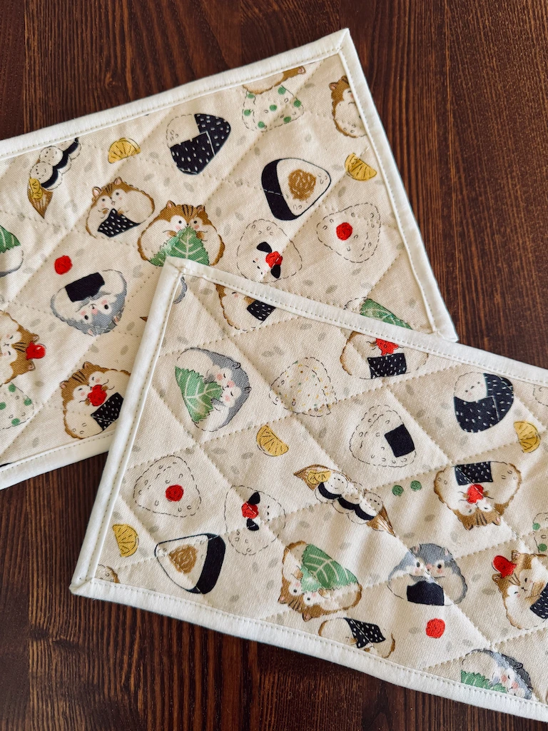 Two 6x9” quilted mug rugs, with stitch lines in a diamond pattern. The cream fabric features hamsters shaped like onigiri and holding items that are usually paired with the rice balls, such as shiso leaves, seaweed, and octopus