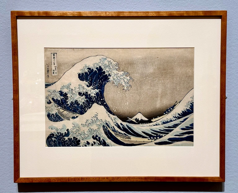 A wood block print of a great tsunami rising up in front of two long boats manned by comparatively small people. Mt Fuji rises far off in the distant background. The block print is in a simple wooden frame.