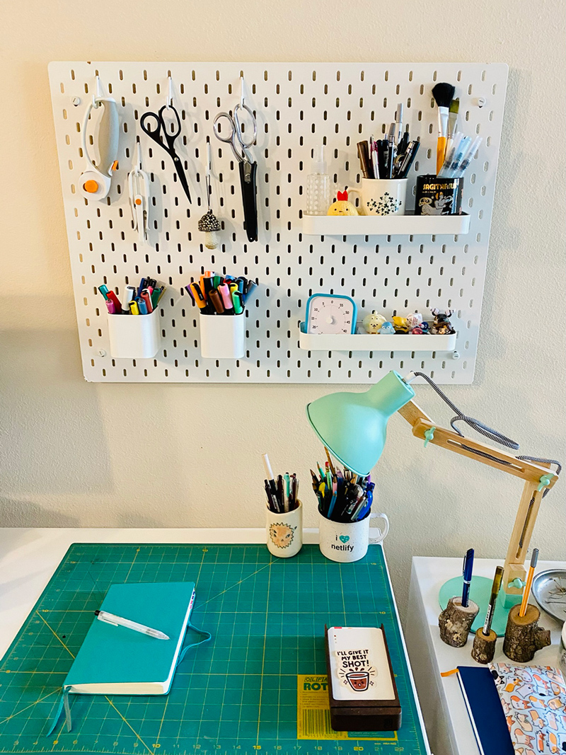 A white pegboard hung above a desk. On the pegboard are hooks bearing scissors and rotary cutters; jars of pencils, pens, and brushes; and a couple shelves holding little figurines and a Pomodoro clock.