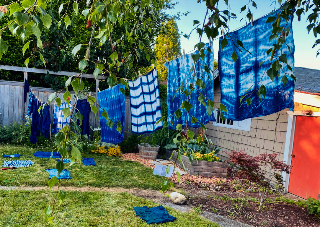 Indigo dyed fabrics hanging on a line in a sunny back yard. Some of the blue and white patterns are strongly graphic while others have loose waves or solid color