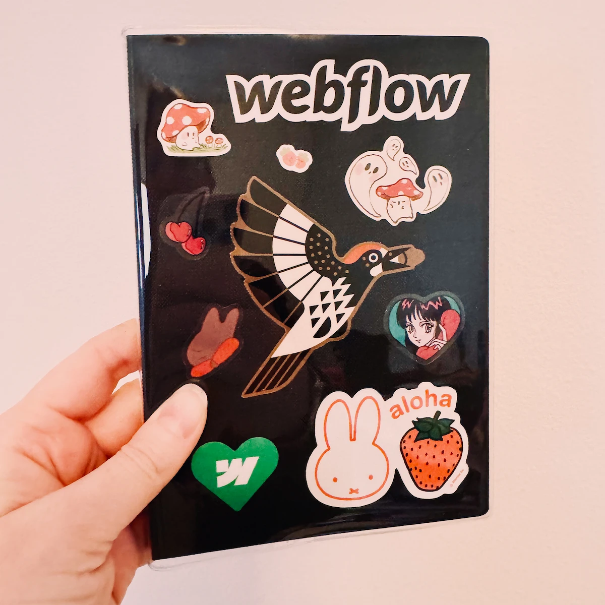 A black journal inside a clear plastic cover. Stuck to the journal itself are various stickers in black, white, red, and green. Their themes include Webflow, mushrooms, Miffy the bunny saying aloha, and other cute aesthetics.