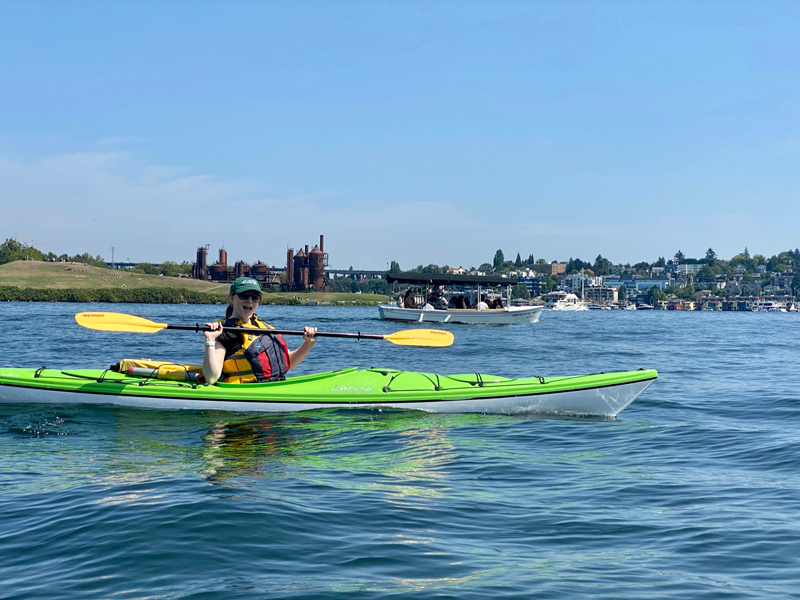 Me sitting in a neon-green kayak, holding my paddle aloft with an excited expression. I am in the middle of a lake with a cityscape behind me.