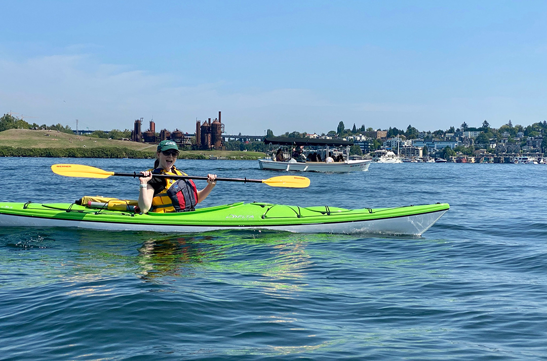 Me sitting in a neon-green kayak, holding my paddle aloft with an excited expression. I am in the middle of a lake with a cityscape behind me.