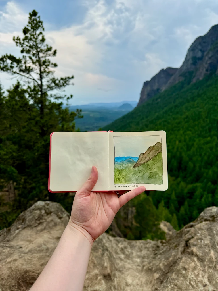 Me holding a small square sketchbook with a watercolor painting of a mountain top, in front of said mountain top