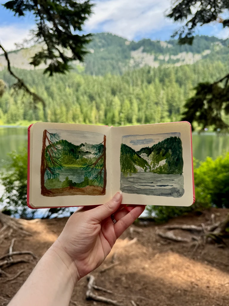 My hand holding the sketchbook in front of another alpine lake. I’ve drawn the two lakes using water color and Neocolor II crayons