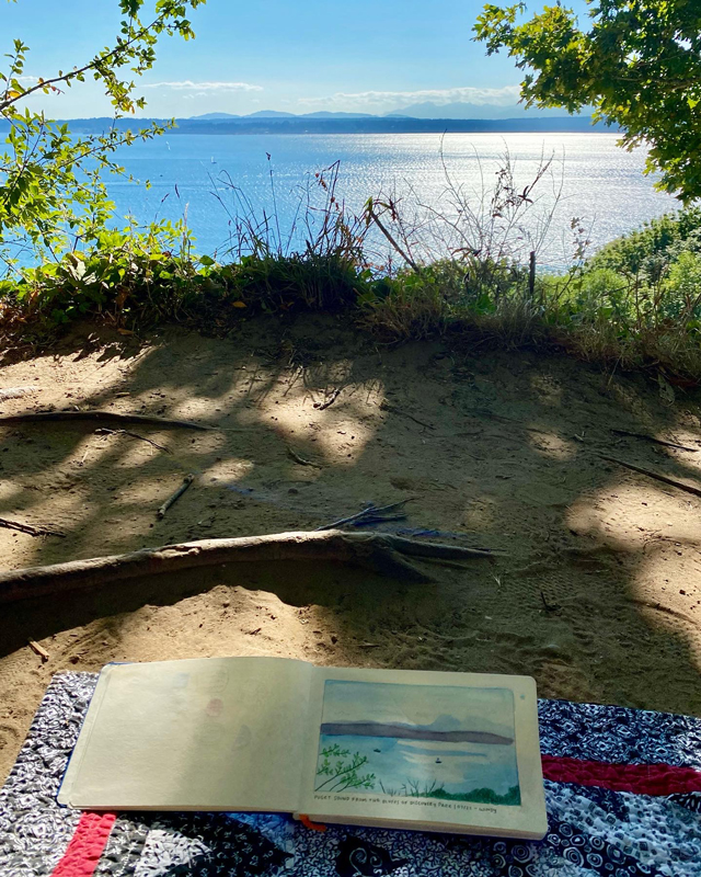 My sketchbook open on a blanket, with a sketch of the water and mountains also pictured beyond.