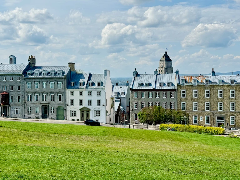 A row of old stone buildings in a French architectural style sitting beyond a bright-green grassy hill. Aturret of the Fairmont Le Chateau Frontenac peeks up in the back.