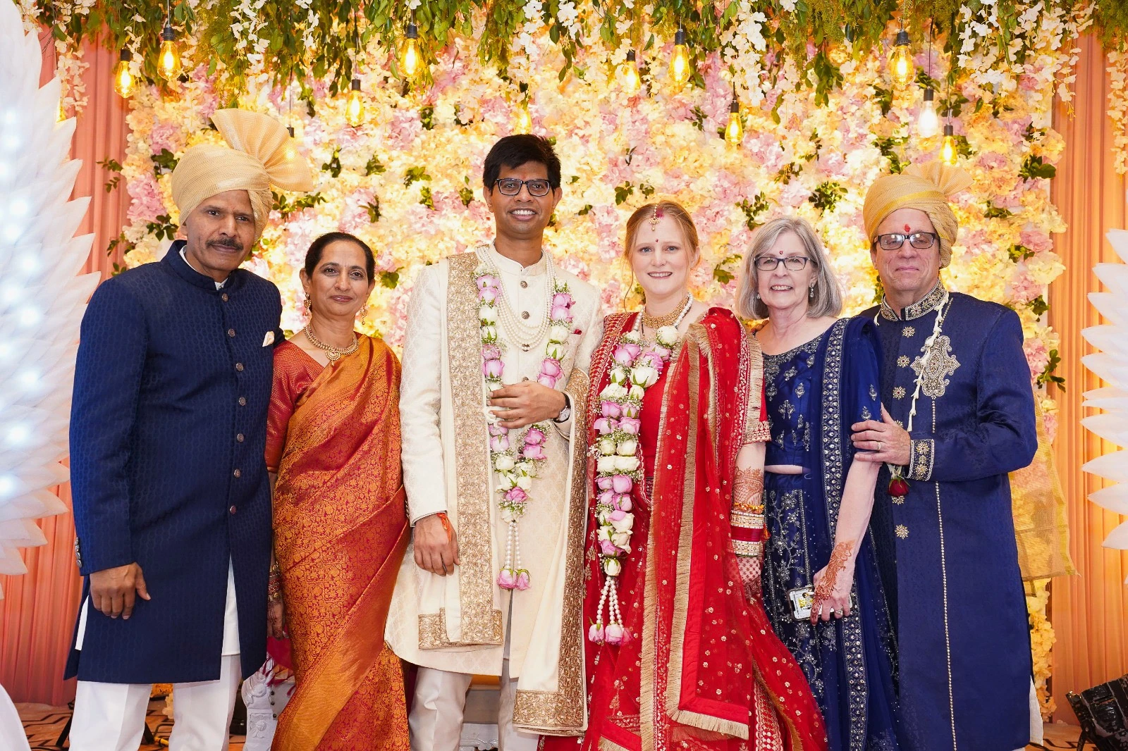 Rahul and I with our parents in traditional Indian wedding garb, in front of a wall full of flowers. I am wearing a bright red lehenga dress. Rahul is wearing a cream-colored suit called a sherwani. Rahul and I both have garlands of pink and yellow flowers around our necks.