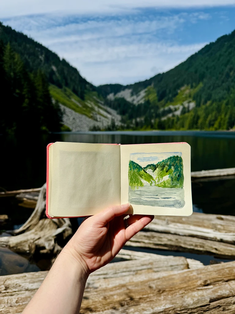 An alpine lake with hills behind, covered in evergreen trees. In the foreground of the lake lie floating logs. In front of this scene, my hand is holding a small square sketchbook with a rendering of the lake.