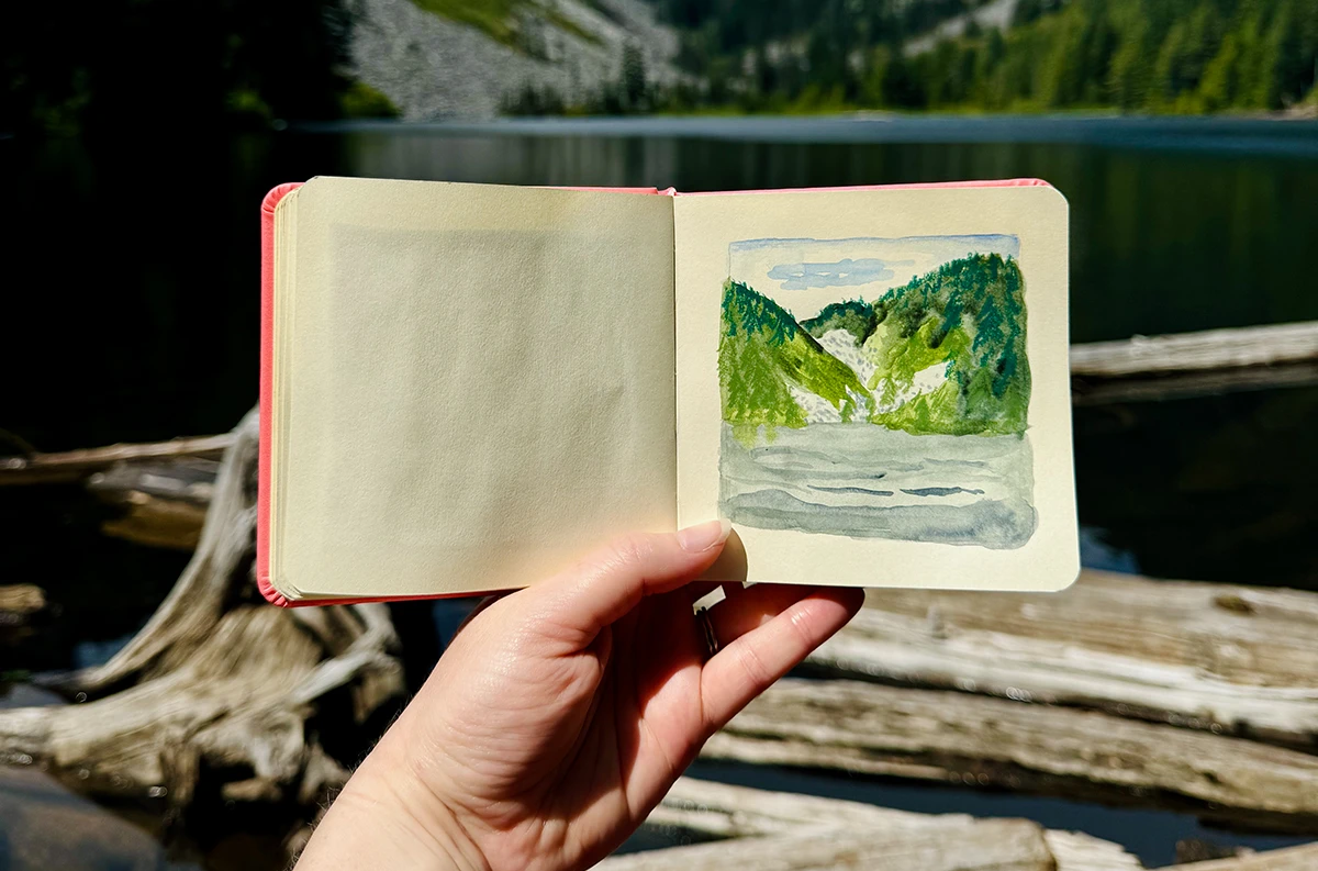 An alpine lake with hills behind, covered in evergreen trees. In the foreground of the lake lie floating logs. In front of this scene, my hand is holding a small square sketchbook with a rendering of the lake.