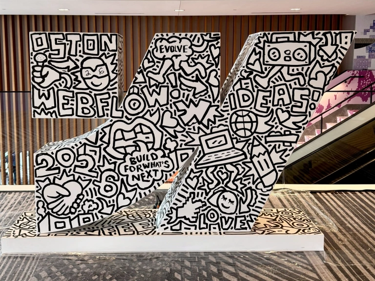 A large white sculpture of the Webflow w mark, covered in conference-themed doodles in black Posca marker