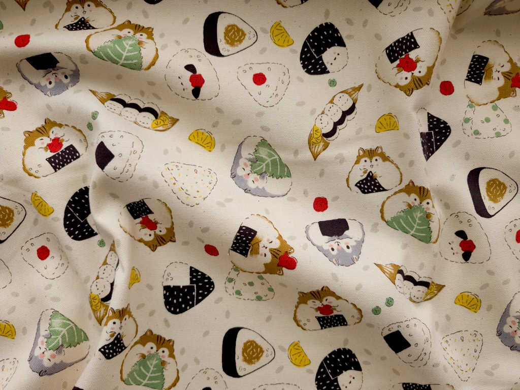A cream fabric with a pattern of illustrated hamsters in triangular shapes, like onigiri. They're holding up things like a shiso leaf or a strip of nori.