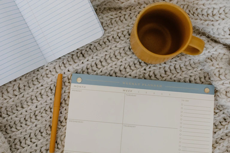 A cozy flatlay of a weekly calendar, pen, notebook, and coffee mug on a thick knitted blanket